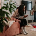 Teen girl uses smartphone while mother stands in colorful, cozy room.