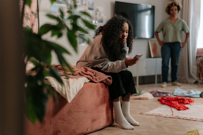 Teen girl uses smartphone while mother stands in colorful, cozy room.