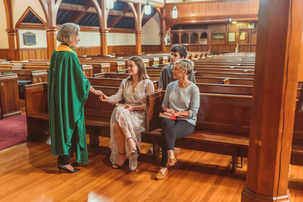 A priest in green chasuble interacts with churchgoers inside a wooden church.